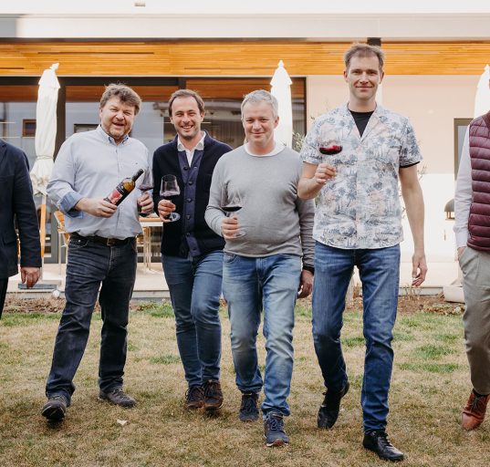 Die vier Winzer und Weintourismus-Obmann Herbert Oschep sowie Weinbaupräsident Andreas Liegegeld mit einem Glas Rotwein in der Han auf einer Wiese.
