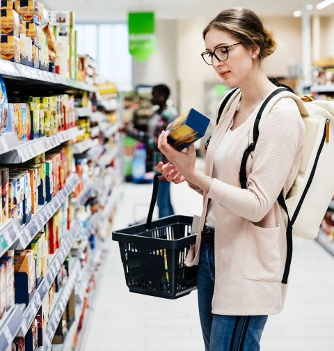 Frau mit Brille, Knoten und rosaroter Weste in Supermarkt mit Korb in der einen und Produkt in der anderen Hand.