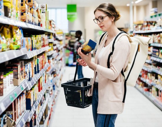 Frau mit Brille, Knoten und rosaroter Weste in Supermarkt mit Korb in der einen und Produkt in der anderen Hand.