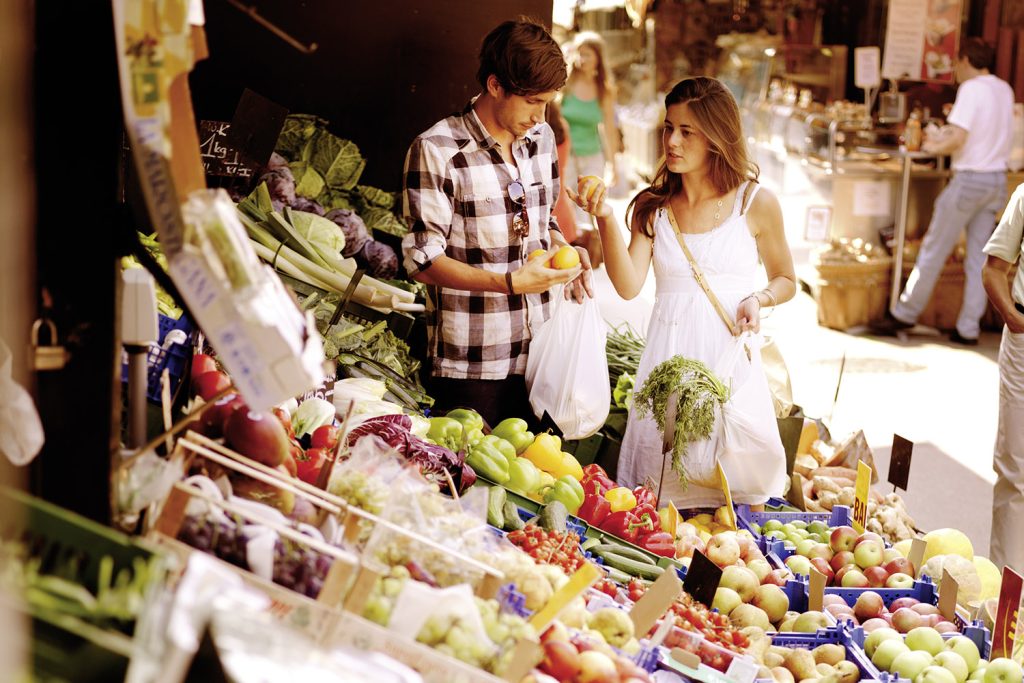 Frau und Mann stehen bei Obst- und Gemüsestand und kaufen ein.