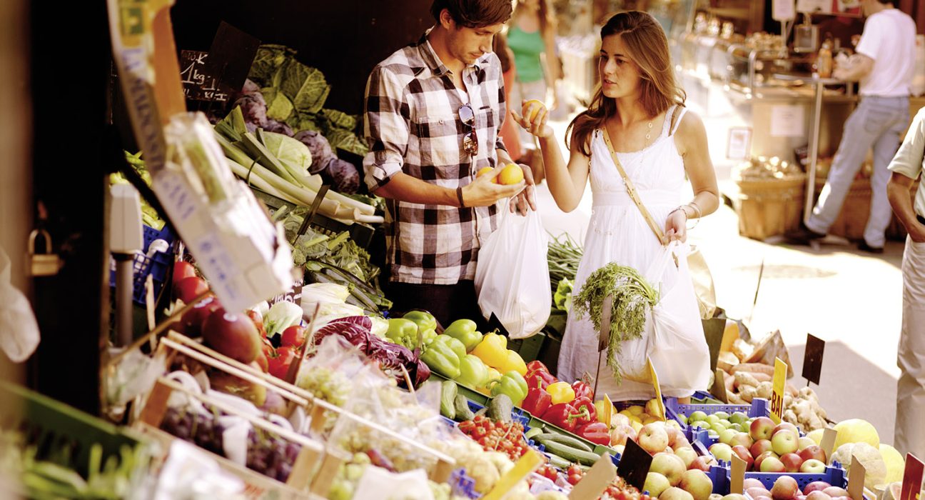 Frau und Mann stehen bei Obst- und Gemüsestand und kaufen ein.