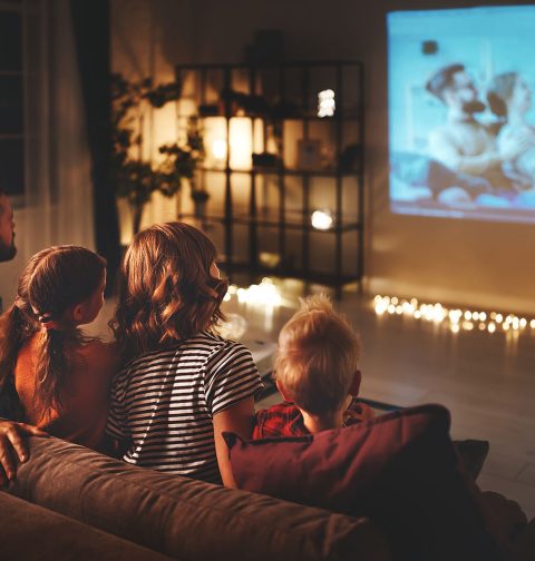 Vater, Mutter, Tochter und Sohn sehen abends auf der Couch über eine Beamer-Leinwand fern. Im Hintergrund sanftes, indirektes Licht und Lichterketten am Boden.