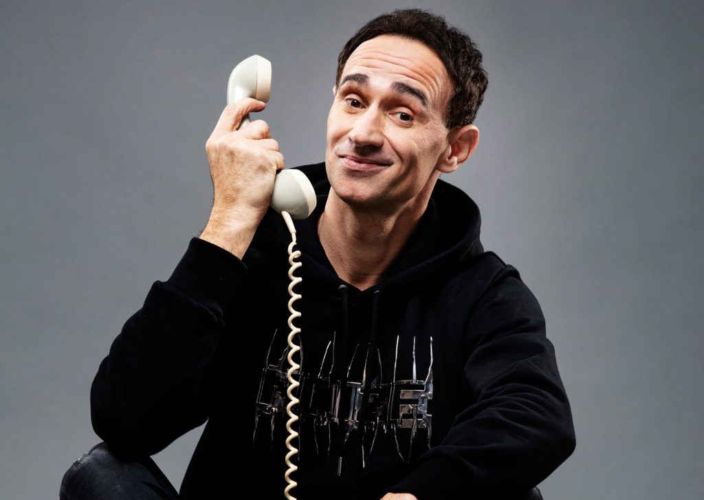 Ö3-Callboy Gernot Kulis mit Telefon in der Hand vor einem grauen Hintergrund.