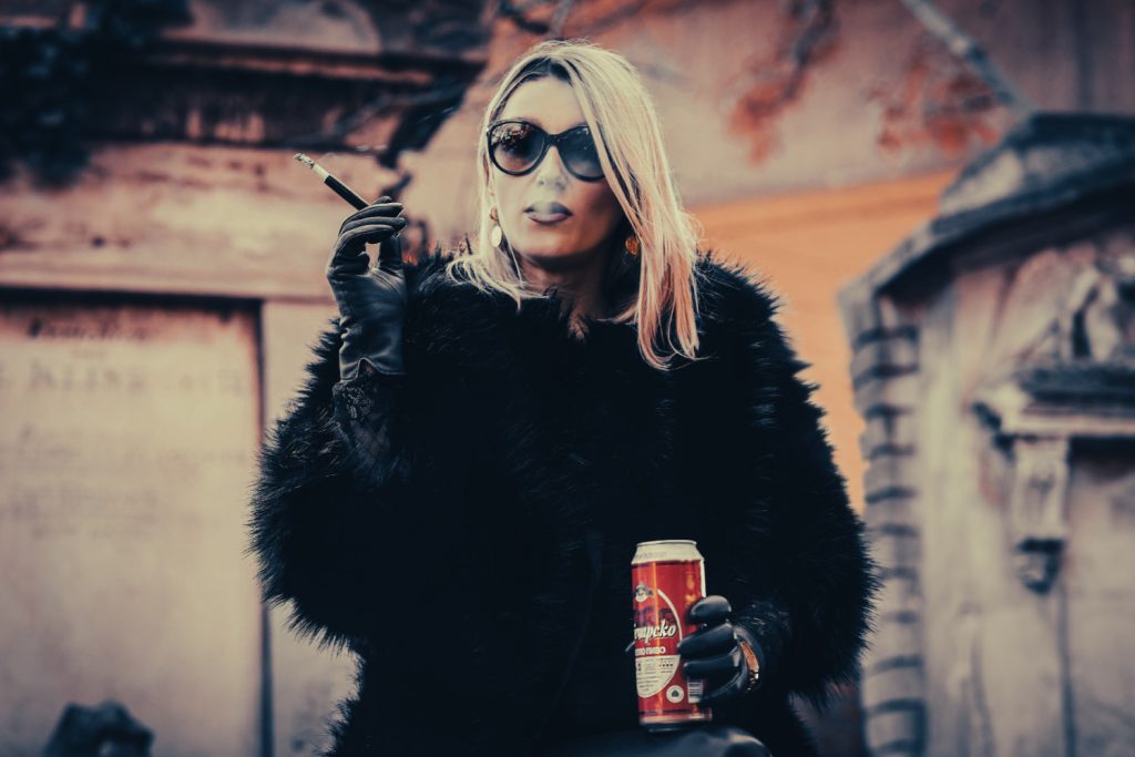 Kabarettistin Malarina steht vor einer gelben Hausmauer mit einer Zigarette und einem Bier in der Hand. Sie trägt eine große schwarze Sonnenbrille, schwarze Lederhandschuhe, eine schwarze Felljacke und eine schwarze lange Lederhose.