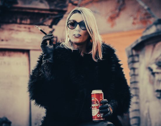Kabarettistin Malarina steht vor einer gelben Hausmauer mit einer Zigarette und einem Bier in der Hand. Sie trägt eine große schwarze Sonnenbrille, schwarze Lederhandschuhe, eine schwarze Felljacke und eine schwarze lange Lederhose.