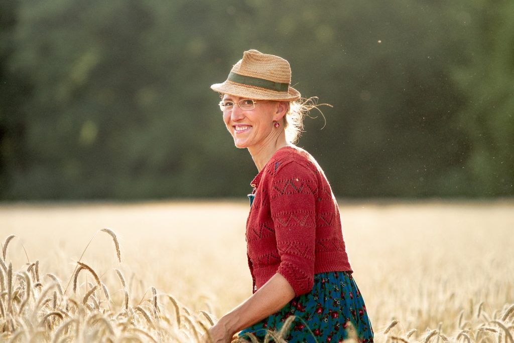 Martina Schwarzmann lächelnd auf einem Getreidefeld vor einem Wald.