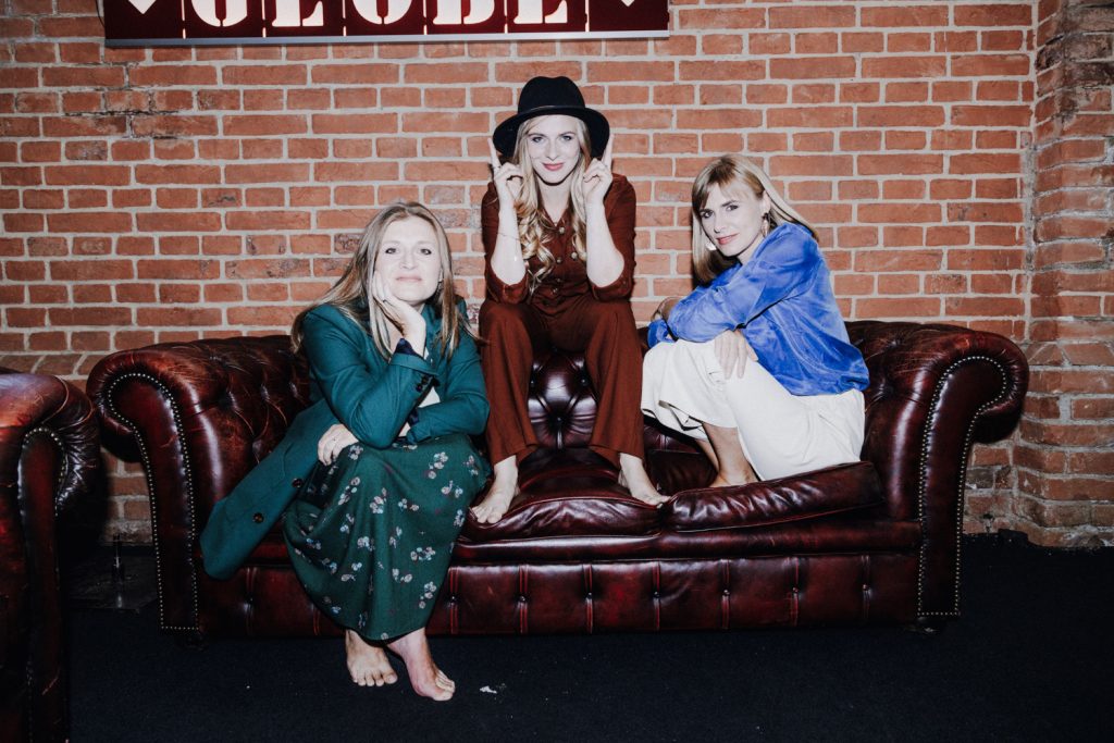 Die Gruppe Poxrucker Sisters auf einer braunen Ledercouch sitzend, vor einer Backsteinziegelwand.