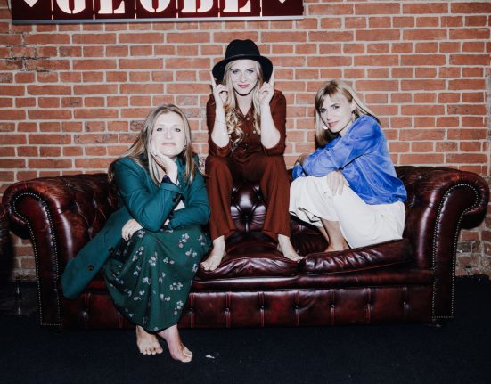 Die Gruppe Poxrucker Sisters auf einer braunen Ledercouch sitzend, vor einer Backsteinziegelwand.
