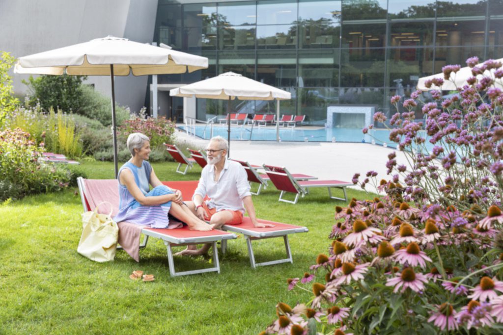 Älteres Pärchen im Außenbereich der Therme Wien unter einem Sonnenschirm auf einer Liege neben purpurnen Sonnenhüten. Im Hintergrund ist eine Glasfront und ein Pool zu sehen.