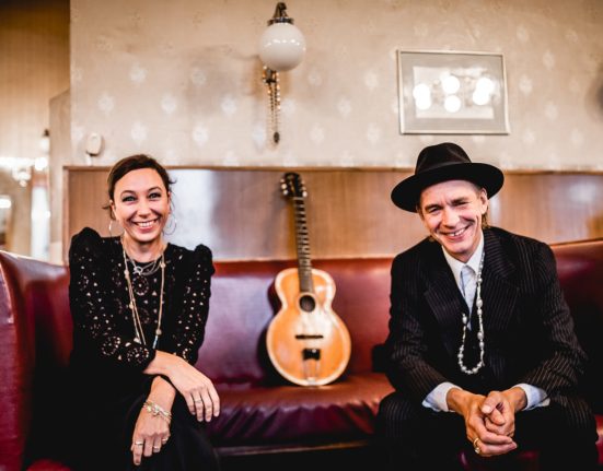 Die Schauspielerin Ursula Strauss und der Songwriter Ernst Molden sitzen auf einer dunkelroten Ledersofa und in der Mitte steht eine Gitarre.