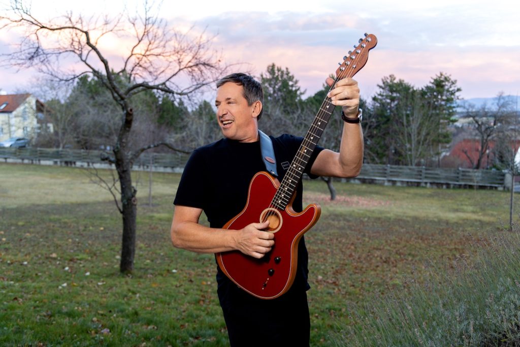 Viktor Gernot steht mit seiner Gitarre auf einer Wiese am Land, hinter ihm Bäume und Häuser. Sein neues Programm heißt „Schiefliegen“.