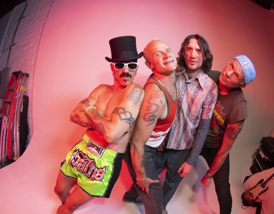 Die vier Musiker der Red Hot Chili Peppers vor einer weißen Wand, rechts sieht man Musikinstrumente.