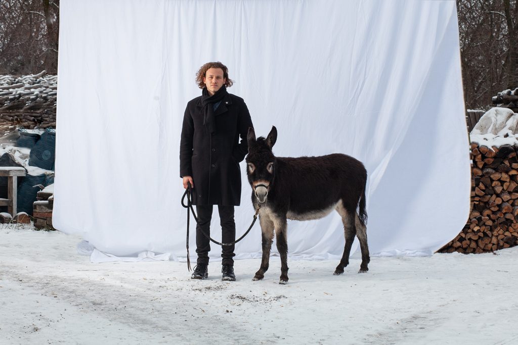 Sänger Cley Freude mit einem Esel in einem schneebedeckten Hof vor einem weißen Laken mit einem Esel an einem Führstrick.
