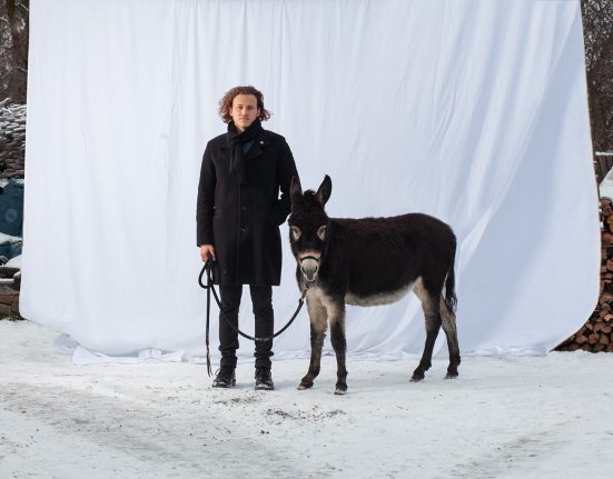 Sänger Cley Freude mit einem Esel in einem schneebedeckten Hof vor einem weißen Laken mit einem Esel an einem Führstrick.