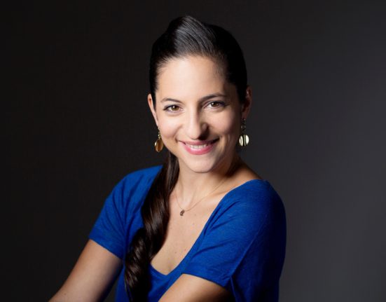 Caroline Athanasiadis mit blauem T-Shirt und verschränkten Armen vor einer grauen Wand.