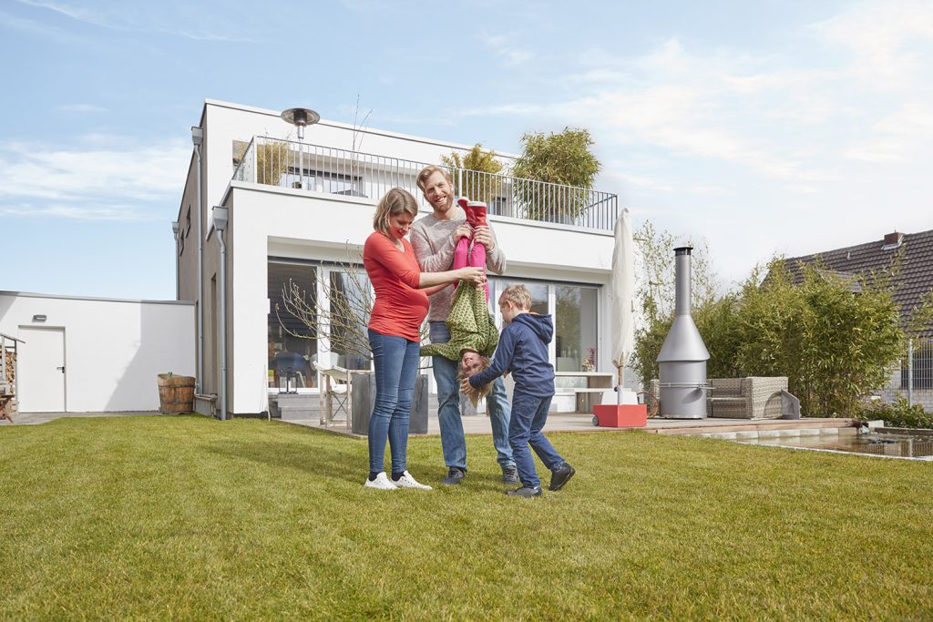 Schwangere Frau und Mann tollen mit zwei Kindern auf grüner Rasenfläche vor einem einstöckigem weißen Haus mit Balkon und Terrasse.