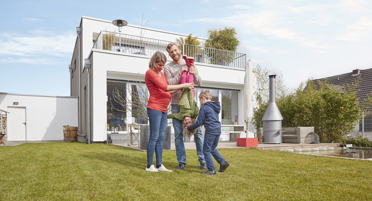 Schwangere Frau und Mann tollen mit zwei Kindern auf grüner Rasenfläche vor einem einstöckigem weißen Haus mit Balkon und Terrasse.