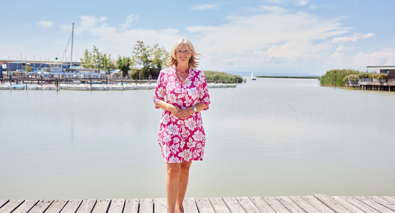 Bürgermeisterin Elisabeth Böhm im wunderschönen Hafen ihrer Gemeinde. Im Hintergrund sieht man Stege, Boote, Schilf und einen azurblauen Himmel mit einigen weißen Wolken.