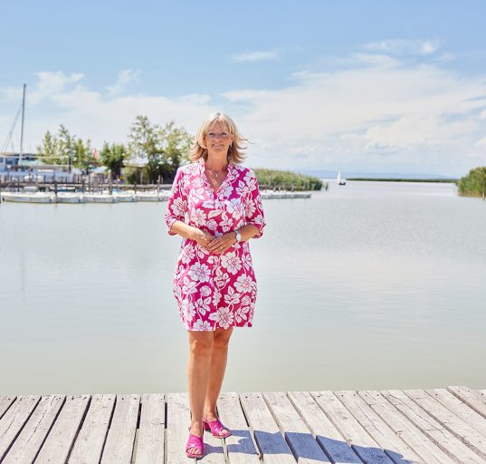 Bürgermeisterin Elisabeth Böhm im wunderschönen Hafen ihrer Gemeinde. Im Hintergrund sieht man Stege, Boote, Schilf und einen azurblauen Himmel mit einigen weißen Wolken.