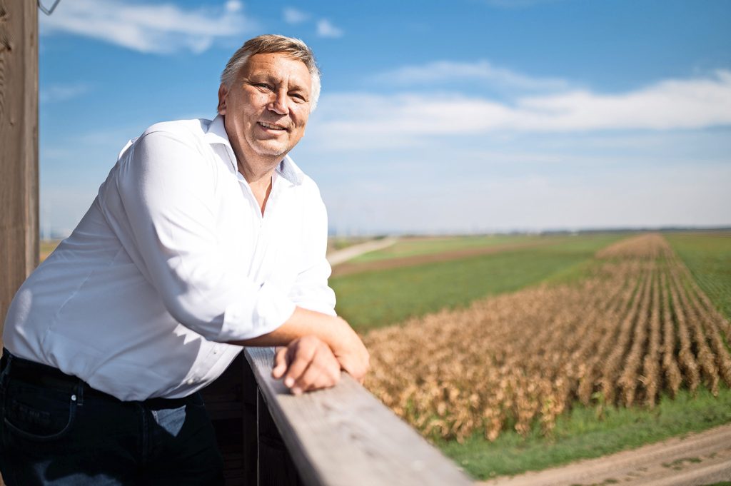 Bürgermeister Manfred Bugnyar in einem hellen Hemd lehnend über ein Geländer. Im Hintergrund sieht man verschwommen Maisfelder und den weiten Himmel.