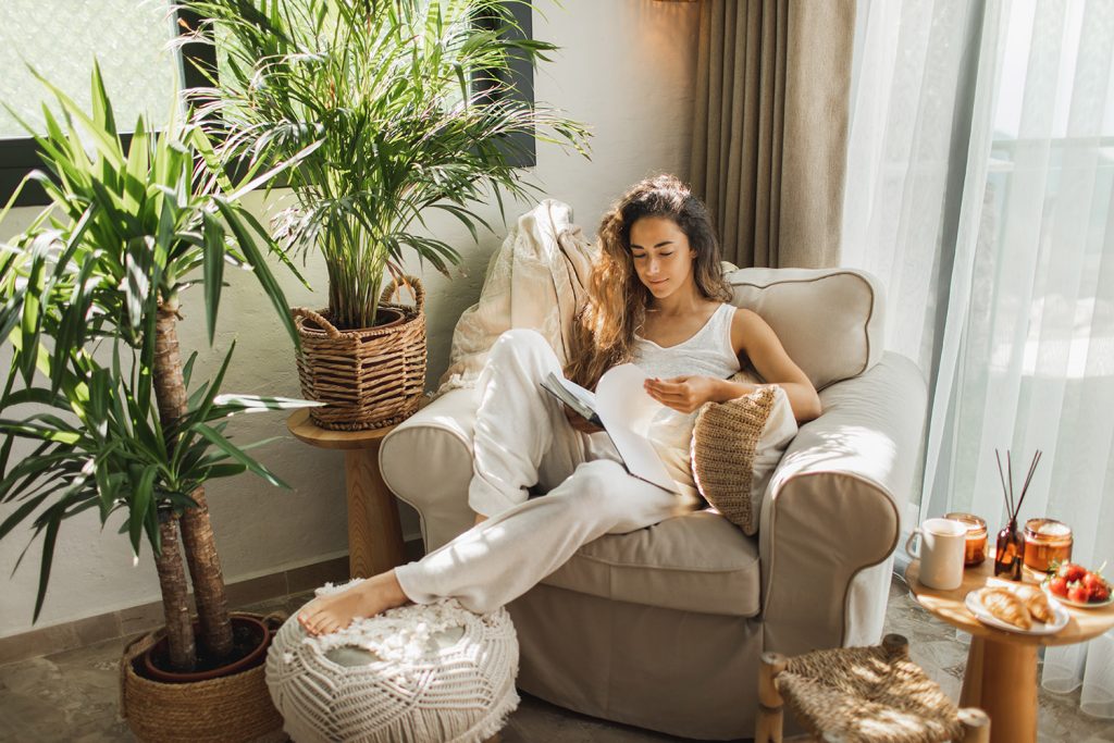 Frau liest zu Hause ein Buch und sitzt dabei auf einem gemütlichen Sessel neben Pflanzen