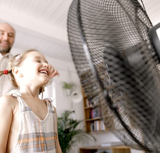 Kind steht vor Ventilator und genieß den Luftzug, hinter ihr steht ihr Vater