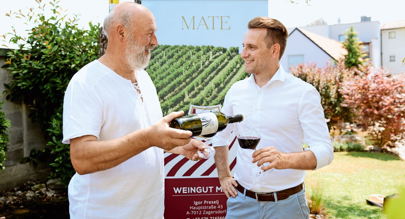 Winzer Mate in kurzärmeligen T-Shirt schenkt Bürgermeister Ivan Grujic Rotwein in Glas ein.