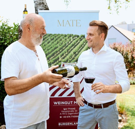 Winzer Mate in kurzärmeligen T-Shirt schenkt Bürgermeister Ivan Grujic Rotwein in Glas ein.