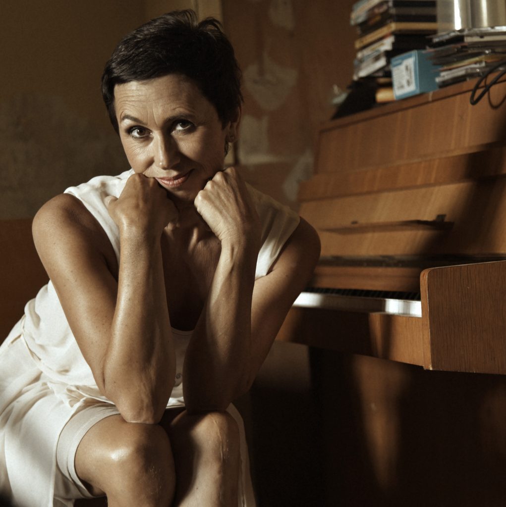 Sängerin Susanne Hell in weißem Kleid mit aufgestützten Armen auf den Knien neben einem Klavier. Auf dem Klavier sind CD-Hüllen zu sehen.