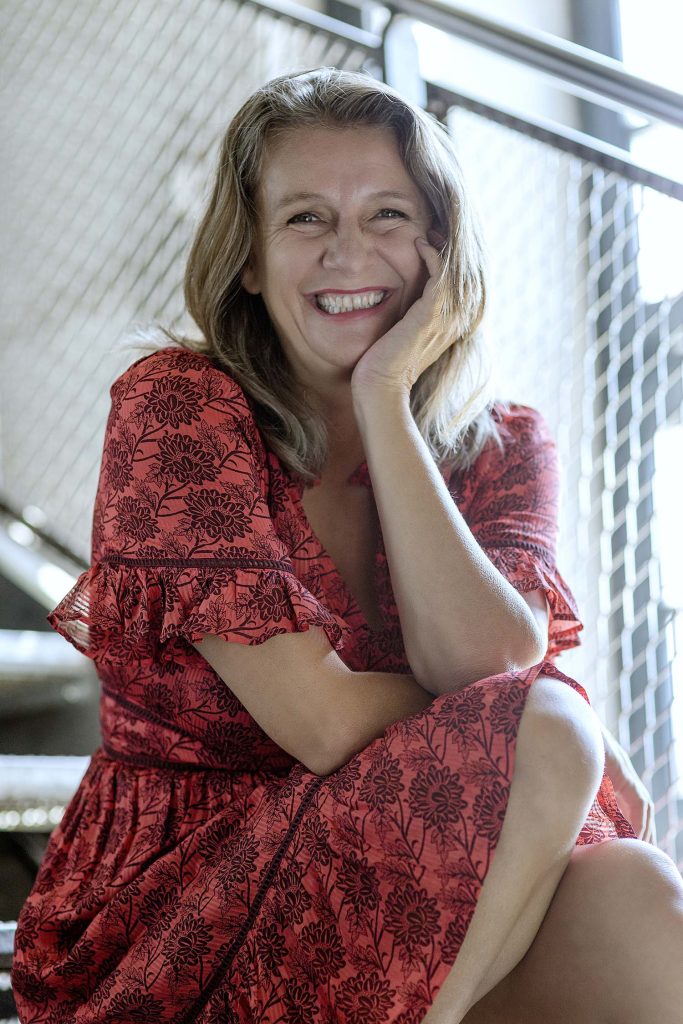 Birgit Denk in rotem Blumenkleid mit kurzen Ärmeln und breitem Lächeln auf dem Gesicht. Sie sitzt auf einer Stiege.