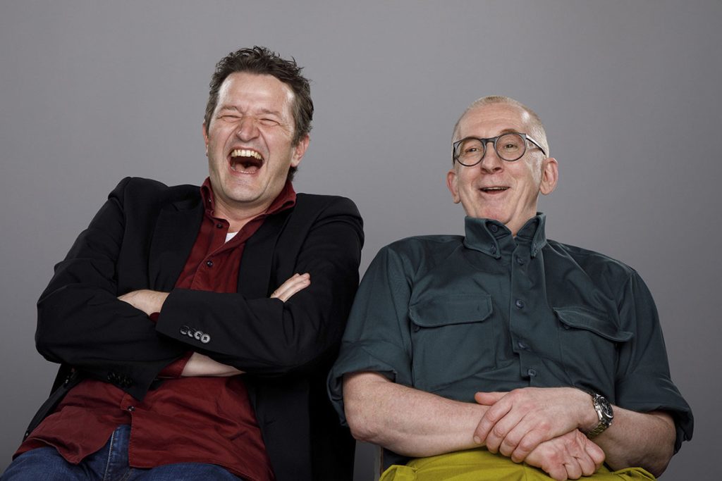 Das Kabarettduo Gunkl und Walter lachend vor einem grauen Hintergrund.