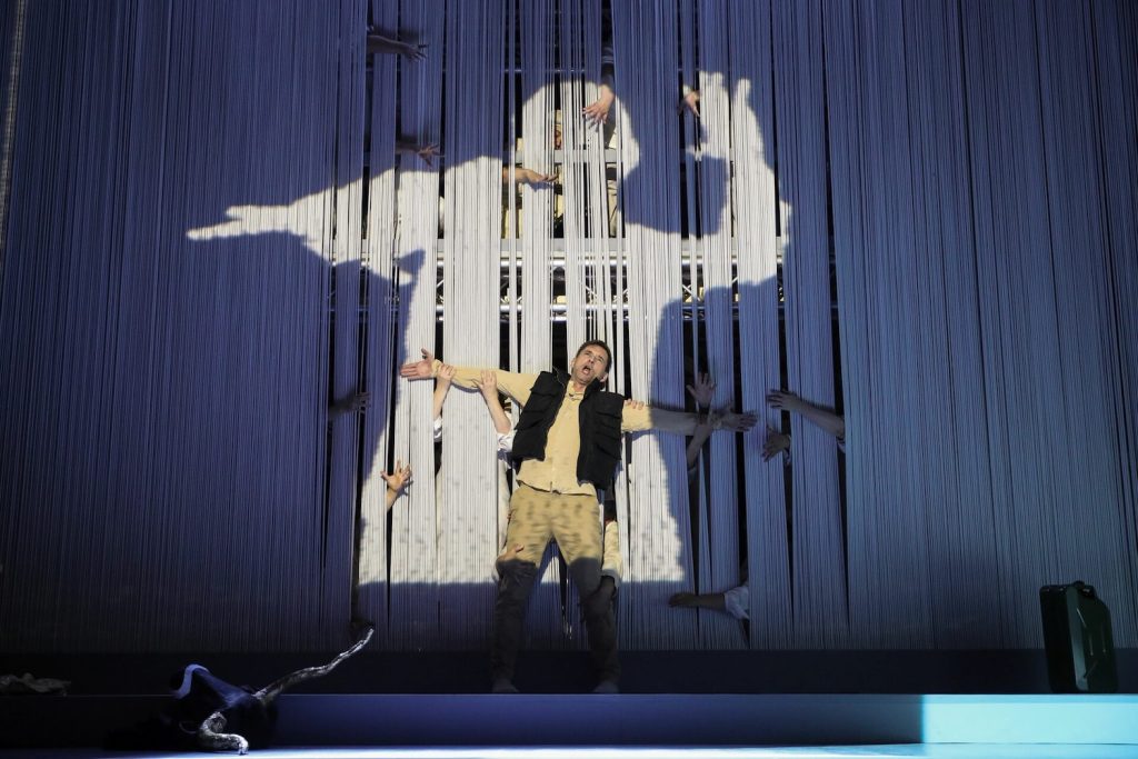 Ein Mann auf einer Bühne vor einem Vorhang, aus dem Hände hervorkommen. Auf dem Vorhang ist auch eine Figur projiziert. © Barbara Palffy