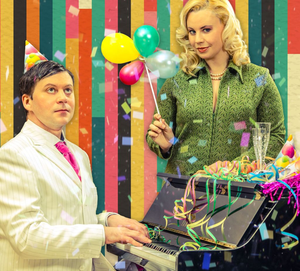 Schauspielerin Katharina Straßer mit Luftballons in der Hand und Boris Fiala, welcher Klavier spielt, verkleidet für eine Geburtstagsparty.