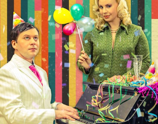 Schauspielerin Katharina Straßer mit Luftballons in der Hand und Boris Fiala, welcher Klavier spielt, verkleidet für eine Geburtstagsparty.