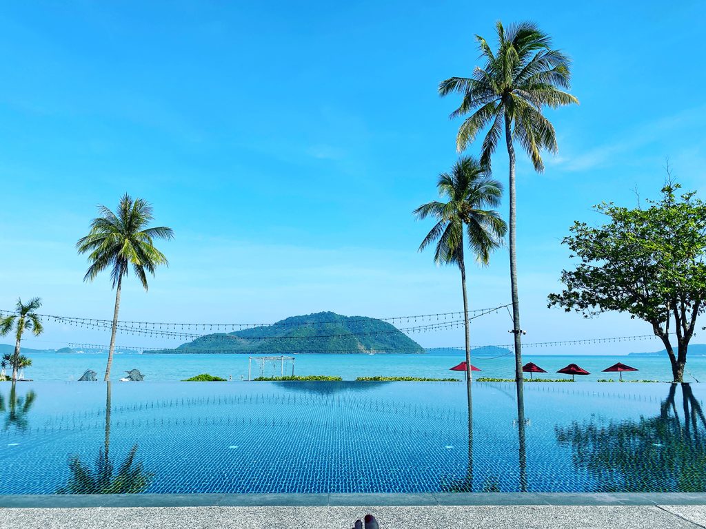 Blick über einen Pool mit Palmen auf das Meer mit einer Insel