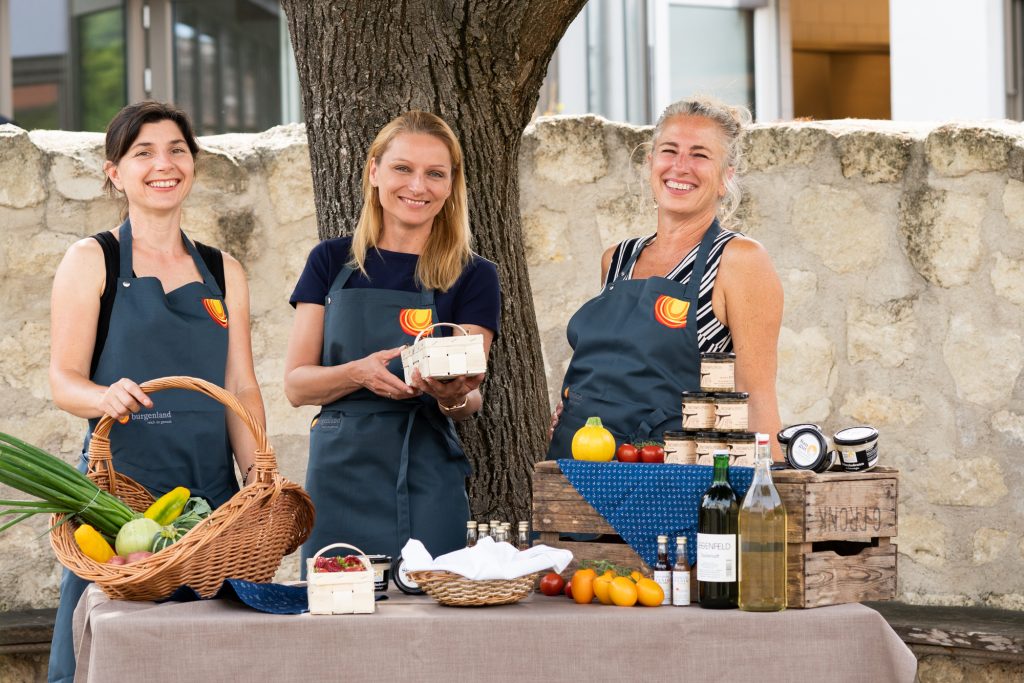 drei Frauen vor einem Tisch mit kulinarischen Spezialitäten lächeln in die Kamera