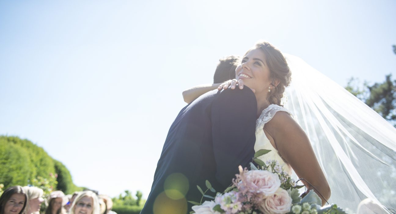 Braut mit Schleier und kurzen Ärmeln umarmt Mann. Ein Blumenstrauß mit hellrosa Rosen befindet sich im Vordergrund. Im Hintergrund sieht man Gäste sitzen und eine grüne Landschaft. Der Himmel ist blau.
