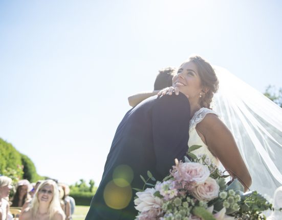 Braut mit Schleier und kurzen Ärmeln umarmt Mann. Ein Blumenstrauß mit hellrosa Rosen befindet sich im Vordergrund. Im Hintergrund sieht man Gäste sitzen und eine grüne Landschaft. Der Himmel ist blau.