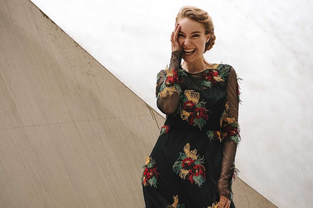 Nikola Hillebrand in einem farbenfrohen Kleid. Sie lacht und hält die rechte Hand an ihr Gesicht. © Miina Jung