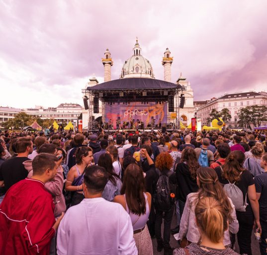 Menschen auf dem Karlsplatz vor einer Bühne. lila Himmel