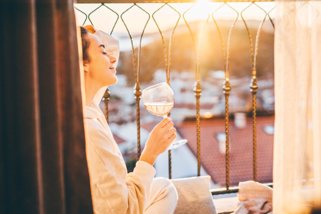 Frau in der Abendsonne auf einer Terrasse mit einem Glas Wein der Hand. Im Vordergrund sind Vorhänge in Sand-Tönen. Im Hintergrund sieht man verschwommen Häuserdächer.