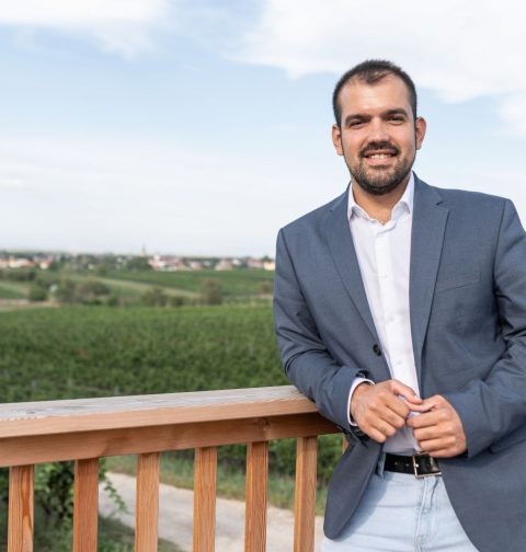 Golser Bürgermeister Kilian Brandstätter lehnt an einer hölzernen Brüstung mit hellen Hemd und grauem Sakko. Im Hintergrund sieht man verschwommen eine grüne Hügellandschaft.