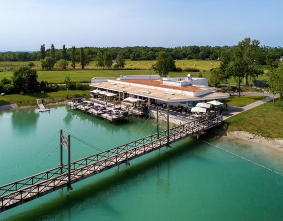 Das Restaurant die Möwe der VILA VITA Pannonia an einem sonnigen Tag. Im Vordergrund der Badesee mit Brücke im Hintergrund sieht man eine grüne Landschaft mit Strohballen und Bäumen.