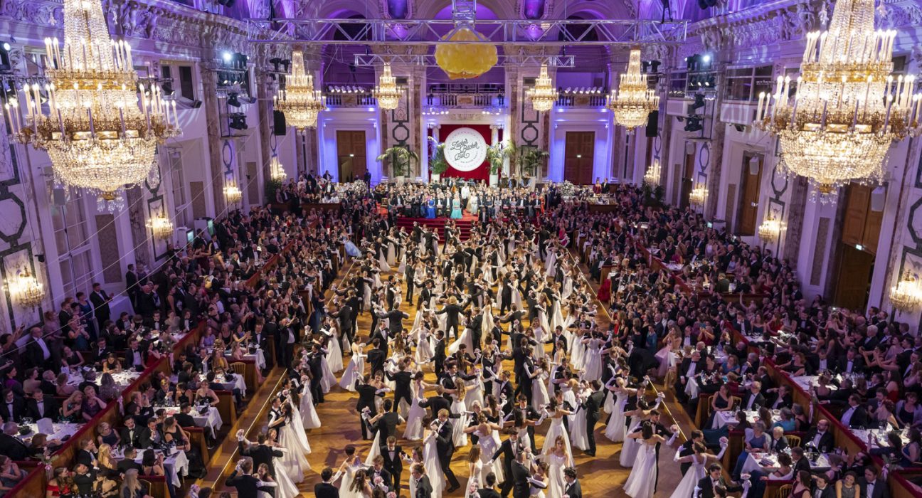 Tänzer und Tänzerinnen im Ballsaal, links und rechts jeweils Tische mit sitzenden Besuchenden in Ballkleidung