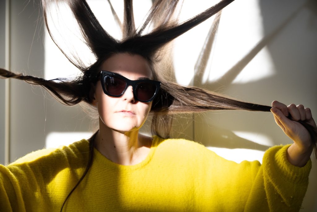 Eine Frau (Doris Uhlich) in gelbem Pullover mit Sonnenbrille, die Haare wie in Strahlen der Sonne von ihrem Kopf abstehend. © Alexi Pelekanos