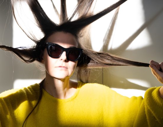 Eine Frau (Doris Uhlich) in gelbem Pullover mit Sonnenbrille, die Haare wie in Strahlen der Sonne von ihrem Kopf abstehend. © Alexi Pelekanos