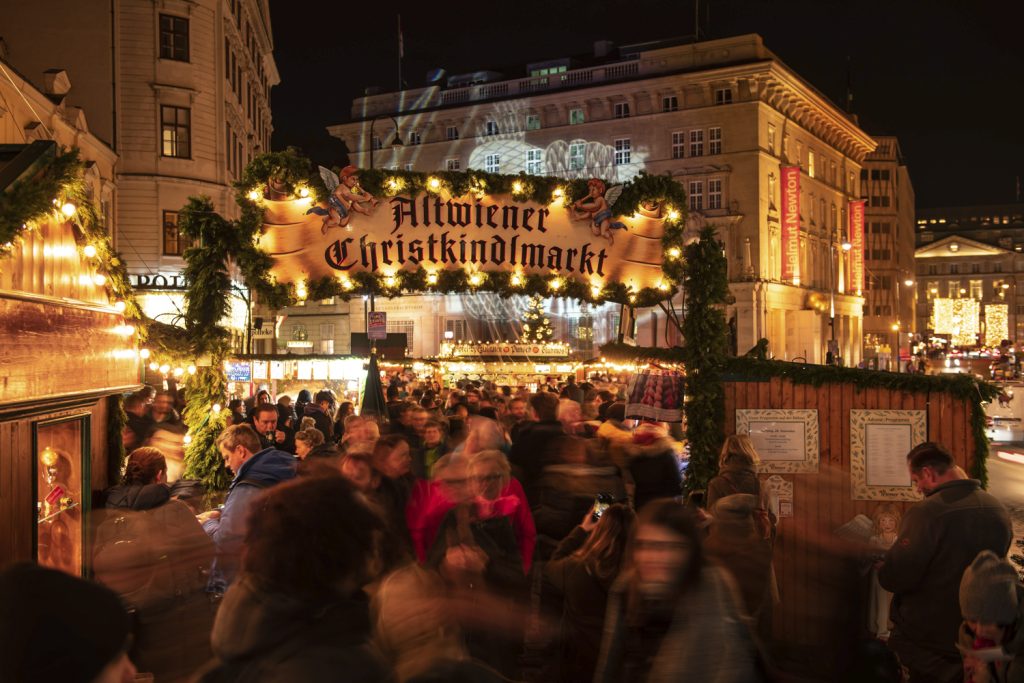 Eingang zum Altwiener Christkindlmarkt auf der Freyung, typische Standl, Menschen in Bewegung, weihnachtliche Deko und Beleuchtung, im Hintergrund das Kunstforum Wien