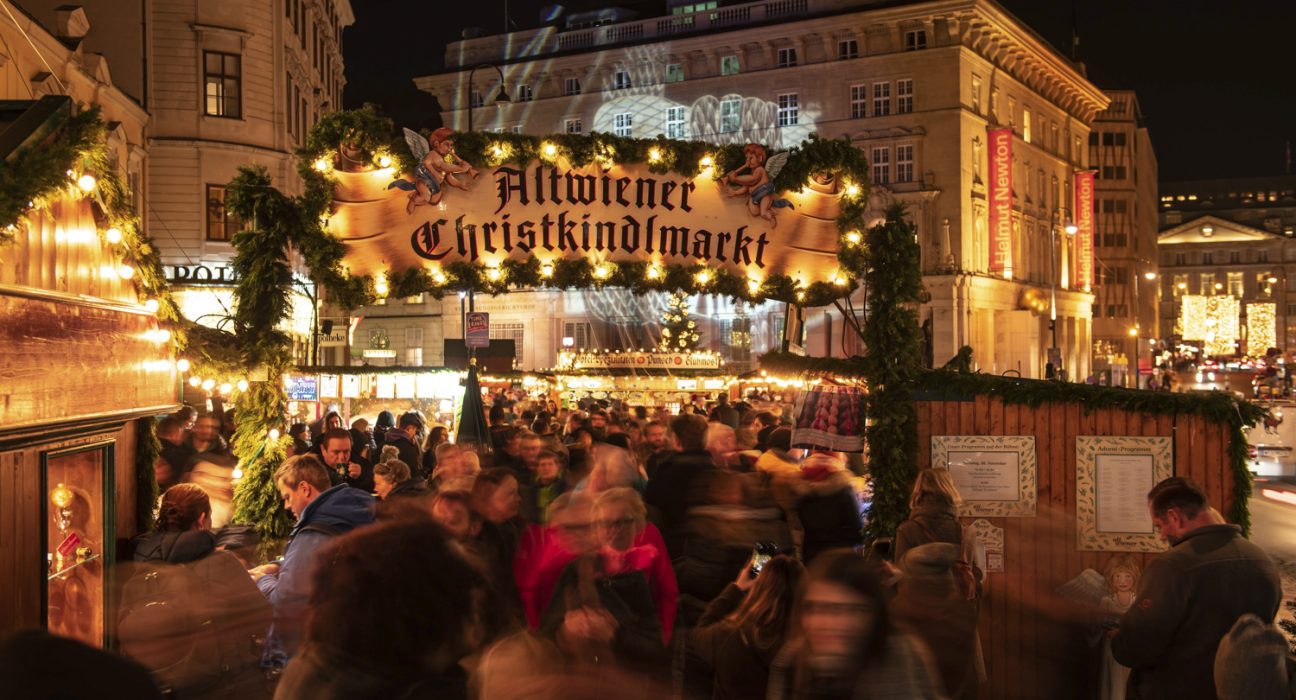 Eingang zum Altwiener Christkindlmarkt auf der Freyung, typische Standl, Menschen in Bewegung, weihnachtliche Deko und Beleuchtung, im Hintergrund das Kunstforum Wien
