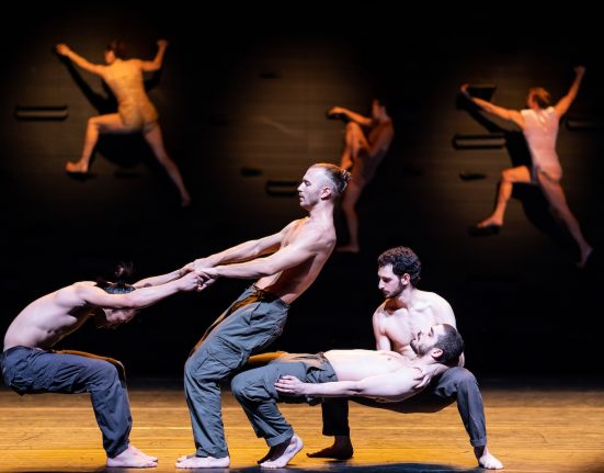Vier Männer mit nackten Oberkörpern in einer Tanzpose auf einer Theaterbühne, im Hintergrund klettern drei Frauen an einer schwarzen Wand hinauf © Ascaf