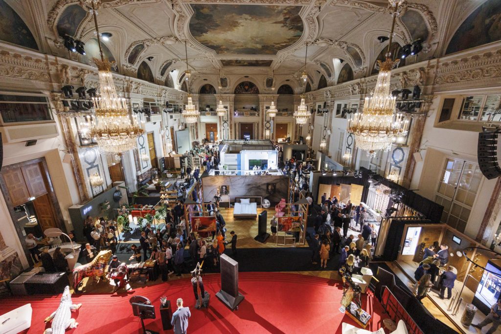 prunkvoller Raum der Hofburg mit Messeständen und Besuchern, Kronleuchter, roter Boden, Gemälde an der Decke